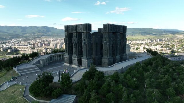 Drone shot capturing the massive bronze and stone pillars of the Chronicle of Georgia monument complex, situated on a green hill overlooking the vast Tbilisi Sea reservoir under a clear blue sky