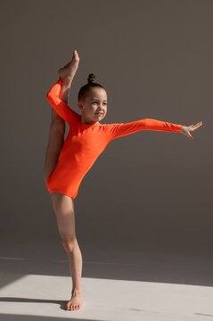 A little Caucasian girl gymnast in an orange leotard demonstrates her skills on a white background. 