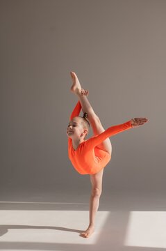 A little Caucasian girl gymnast in an orange leotard demonstrates her skills on a white background. 