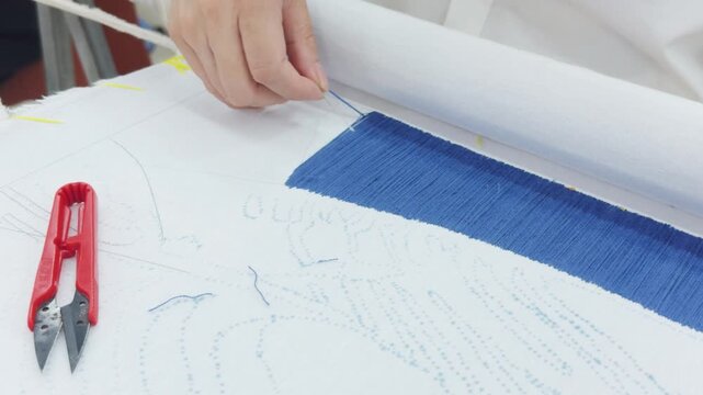 Hand working on a textile piece, a craftsman's hands carefully create intricate embroidery stitches using blue silk thread on white fabric, following a pre drawn pattern