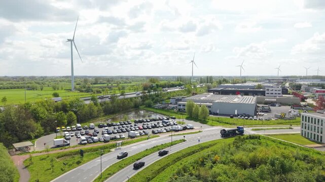 Vertical screen. Aerial perspective of industrial zone featuring parked cars, wind turbines, lush fields, highlighting ongoing movement of vehicles along the road and the surrounding natural scenery