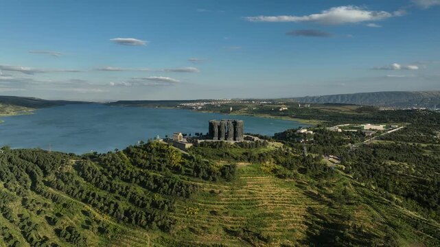 Drone shot capturing the massive bronze and stone pillars of the Chronicle of Georgia monument complex, situated on a green hill overlooking the vast Tbilisi Sea reservoir under a clear blue sky