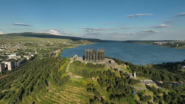 Drone shot capturing the massive bronze and stone pillars of the Chronicle of Georgia monument complex, situated on a green hill overlooking the vast Tbilisi Sea reservoir under a clear blue sky