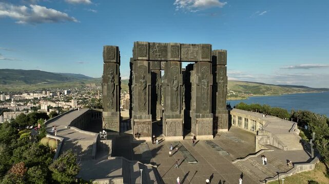 Drone shot capturing the massive bronze and stone pillars of the Chronicle of Georgia monument complex, situated on a green hill overlooking the vast Tbilisi Sea reservoir under a clear blue sky