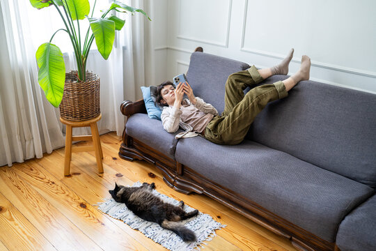 Lazy woman browsing social media on smartphone while lounging on sofa. Zoomer girl wasted time, cat lying on the floor. Device dependency, endless scrolling habit, lonely isolation. Phone addiction. 