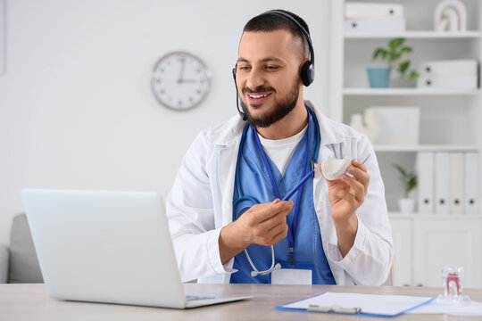 Male dentist with headset and jaw model giving online consultation at table in clinic