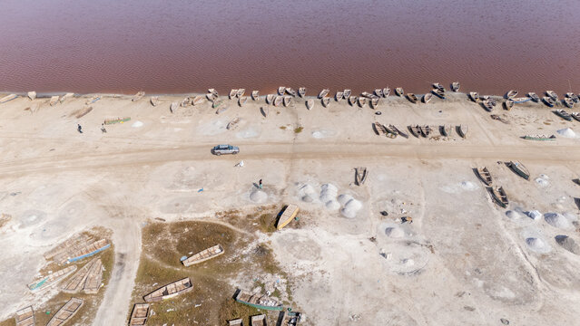 Aerial view of Lake Retba with its distinctive pink water, numerous wooden salt-harvesting boats moored along the sandy shore, and piles of harvested salt in Rufisque, Dakar Region, Senegal.