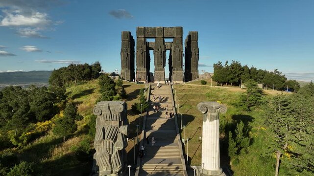 Drone shot capturing the massive bronze and stone pillars of the Chronicle of Georgia monument complex, situated on a green hill overlooking the vast Tbilisi Sea reservoir under a clear blue sky