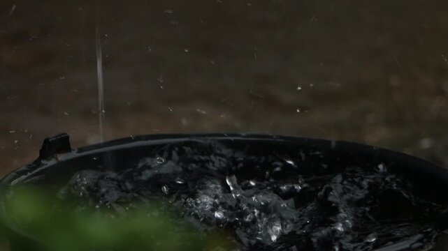 Heavy rain droplets splashing into water collection bucket, close up dramatic water drops falling into full bucket during tropical monsoon rain