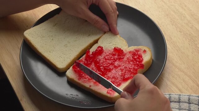 hands using a butter knife to spread bright red strawberry jam onto slices of fresh white bread on a black plate
