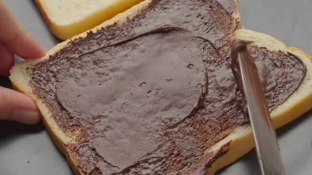 Close up video of hands using a butter knife to spread creamy chocolate hazelnut spread evenly across a slice of white sandwich bread on a black plate
