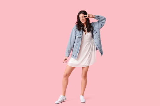 Beautiful young woman in stylish white dress dancing and showing victory gesture on pink background