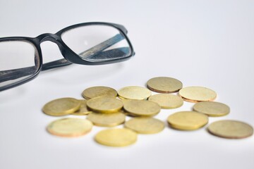 Close-up of a pair of black-framed reading glasses resting next to a scattered heap of gold Malaysian coins on a white background, suggesting financial planning or auditing. © Akram