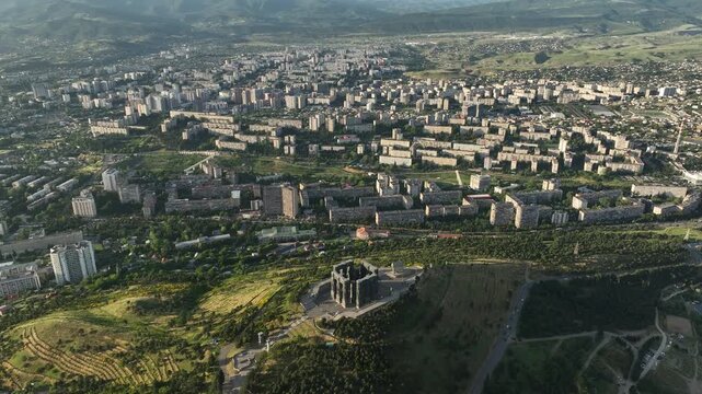 Drone shot capturing the massive bronze and stone pillars of the Chronicle of Georgia monument complex, situated on a green hill overlooking the vast Tbilisi Sea reservoir under a clear blue sky