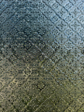 Close-up of patterned, opaque glass featuring a repetitive, embossed floral or cloverleaf design, with light filtering through