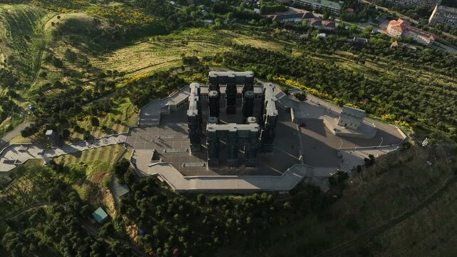 Drone shot capturing the massive bronze and stone pillars of the Chronicle of Georgia monument complex, situated on a green hill overlooking the vast Tbilisi Sea reservoir under a clear blue sky