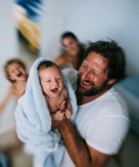 Fototapeta premium Laughing baby wrapped in a blue plush towel held by a happy father for family lifestyle and newborn care concepts