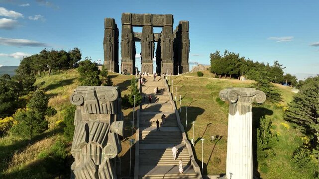Drone shot capturing the massive bronze and stone pillars of the Chronicle of Georgia monument complex, situated on a green hill overlooking the vast Tbilisi Sea reservoir under a clear blue sky