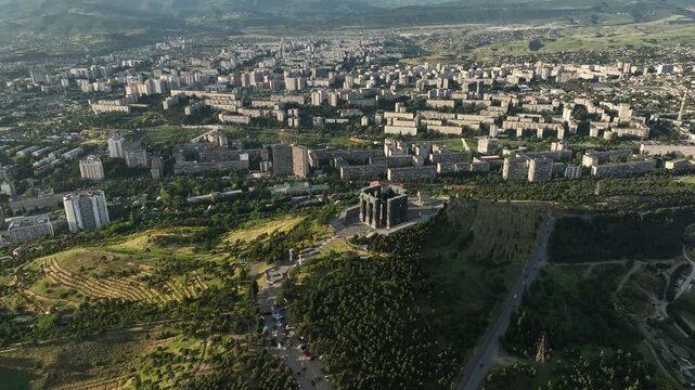 Drone shot capturing the massive bronze and stone pillars of the Chronicle of Georgia monument complex, situated on a green hill overlooking the vast Tbilisi Sea reservoir under a clear blue sky