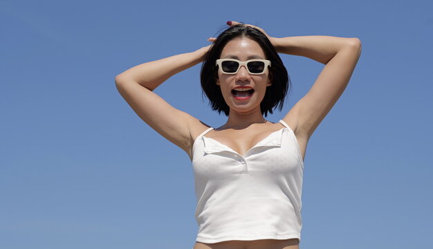 Joyful young Asian woman wearing white sunglasses and a white top posing with her hands on her head against a clear blue sky background