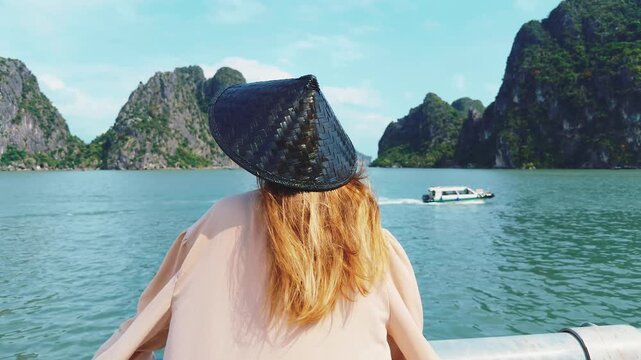 Woman wearing a non la hat looking at the beautiful ha long bay landscape, exploring iconic limestone karsts and the serene sea while traveling by boat in vietnam