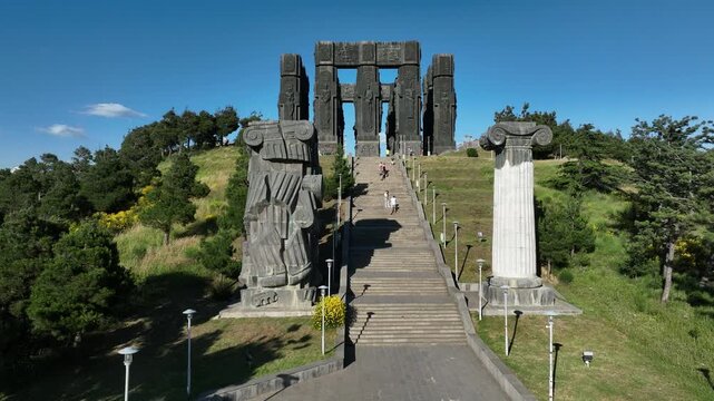 Drone shot capturing the massive bronze and stone pillars of the Chronicle of Georgia monument complex, situated on a green hill overlooking the vast Tbilisi Sea reservoir under a clear blue sky