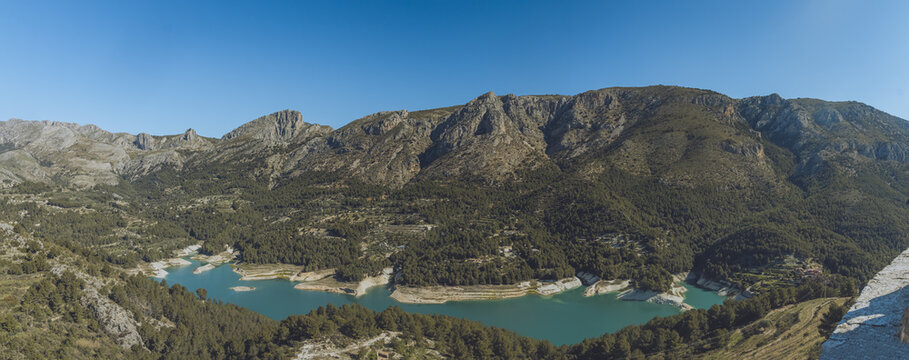 Aerial view of the Guadalest Reservoir surrounded by rugged mountains, pine forests, and rocky terrain in Alicante, Valencian Community, Spain.