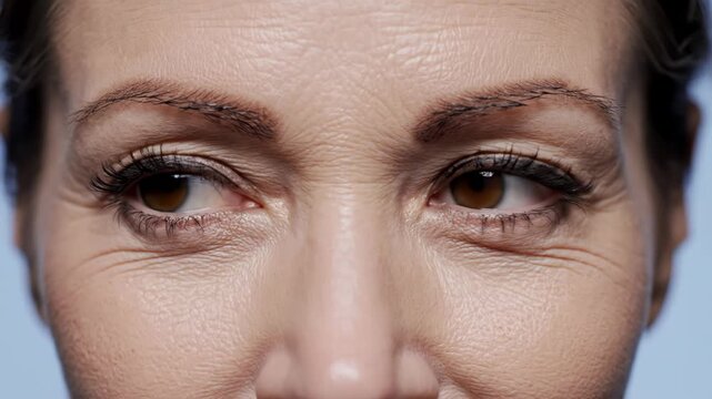 Extreme close-up of mature woman's brown eyes, featuring thick dark eyebrows, black mascara on lashes, and detailed skin texture with wrinkles, crow's feet, and pores on forehead.