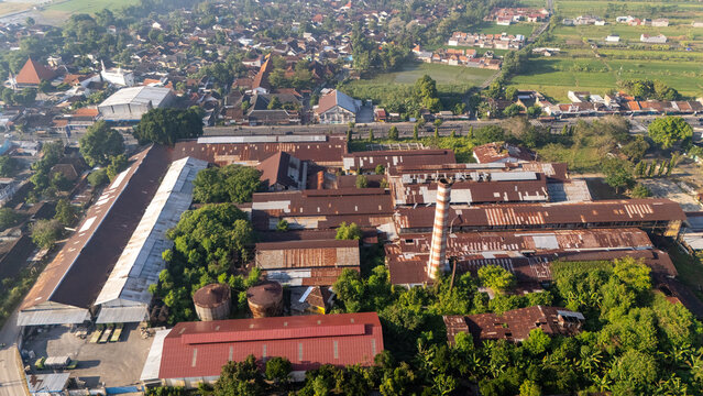 Aerial view of an old industrial factory complex with rusted metal roofs, a tall brick chimney, and surrounding lush greenery in Indonesia.