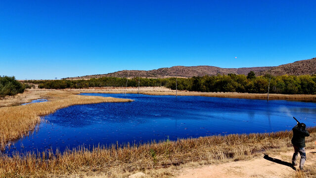 Aerial view of a person shooting a shotgun near a blue pond surrounded by dry grass and hills under a clear blue sky in iNtabazwe, Free State, South Africa.
