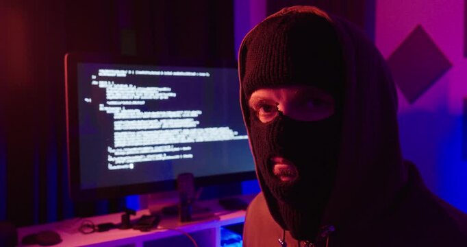 Masked person in a hooded sweatshirt looking toward the camera with a computer monitor displaying code in the background. Concept of cybersecurity, cybercrime, hacking, digital threats, anonymity
