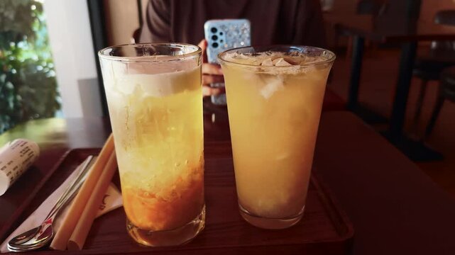 Two tall glasses of cold beverages. Including a layered fruit tea and an iced drink. Sit on a wooden serving tray beside a rolled receipt and bio degradable straws
