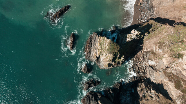 Aerial view of the rugged cliffs and rocky coastline of Burgh Island with turquoise water and crashing waves in Kingsbridge, Devon, United Kingdom.
