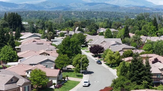 Single-family homes with terracotta rooftops, manicured lawns and tree-lined streets spreading across quiet suburban neighborhood under sunny skies in Redding, California, USA. Aerial drone tilt up