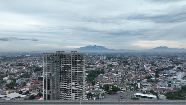 Aerial view of an unfinished high-rise building surrounded by dense urban sprawl and residential rooftops under a cloudy sky in Bekasi Barat, West Java, Indonesia.