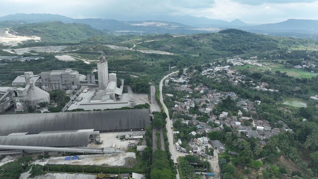 Aerial view of a large cement factory complex situated next to a dense residential neighborhood with lush green hills in the background in Klapanunggal, West Java, Indonesia.