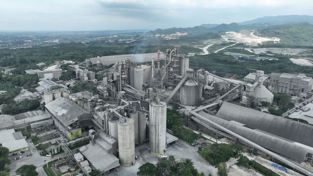 Aerial view of a large industrial cement factory with tall silos, processing structures, and conveyor belts surrounded by green hills in Klapanunggal, West Java, Indonesia.