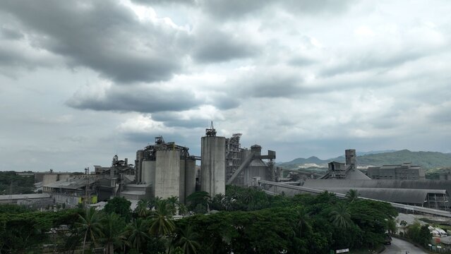 Aerial view of the industrial cement factory with large concrete silos, conveyor systems, and surrounding lush tropical forest under a cloudy sky in Klapanunggal, West Java, Indonesia.