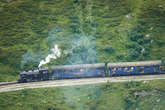 Aerial view of the historic Furka Steam Railway steam locomotive pulling blue passenger carriages through a lush green alpine mountain landscape, Switzerland.