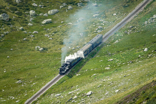 Aerial view of a historic steam train traveling along the tracks of the Dampfbahn Furka Bergstrecke through the green alpine landscape of Switzerland.