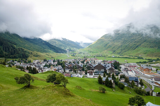Aerial view of the village of Andermatt nestled in the green valley of the Swiss Alps, surrounded by lush meadows, mountain slopes, and cloudy skies in Andermatt, Uri, Switzerland.