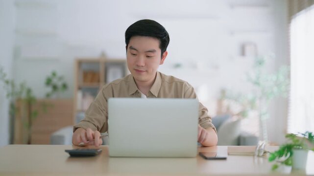 Happy young Asian man working on laptop in living room. Freelance worker male sitting at desk writing notes while watching webinar, studying online