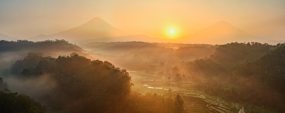Aerial view of lush rice terraces nestled between misty mountains at sunrise in Selomerto, Central Java, Indonesia.