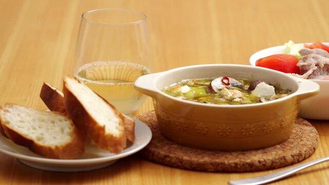 Seafood and mushroom ajillo on a wooden dinner table with white wine, sliced baguette, and salad, close up shot.