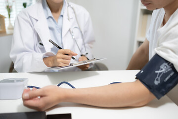 Doctor records patient's blood pressure reading. Patient rests arm with cuff securely fitted. Clinic setting suggests routine health monitoring. Professional demeanor shows care and precision   © Somdetmeaow