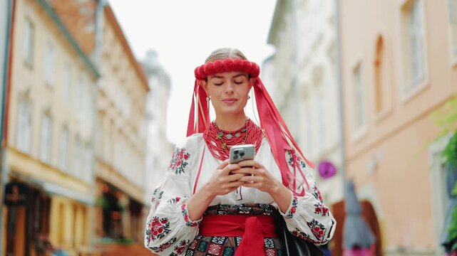 Smiling woman dressed in traditional Ukrainian vyshyvanka and red wreath, typing message and scrolling on smartphone on historic cobblestone street. Urban lifestyle concept.