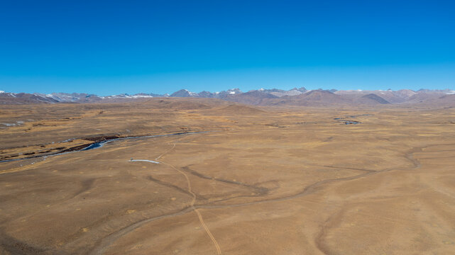Aerial view of Deosai National Park featuring a vast arid plateau, winding river, and distant snow-capped mountains under a clear blue sky in Astore, Gilgit, Pakistan.