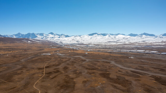 Aerial view of Deosai National Park featuring vast brown plains, winding dirt tracks, and snow-capped mountain peaks under a clear blue sky in Astore, Gilgit, Pakistan.