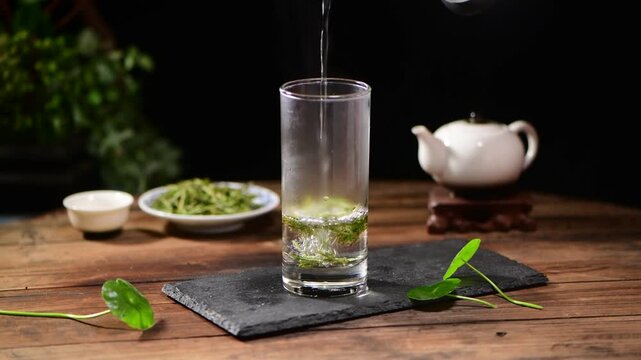 Pouring hot water into a glass to brew green tea on a rustic wooden table with a teapot background