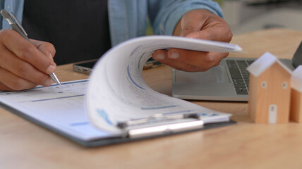 Close up of a homeowner reviewing mortgage documents and signing paperwork at home office. Real...